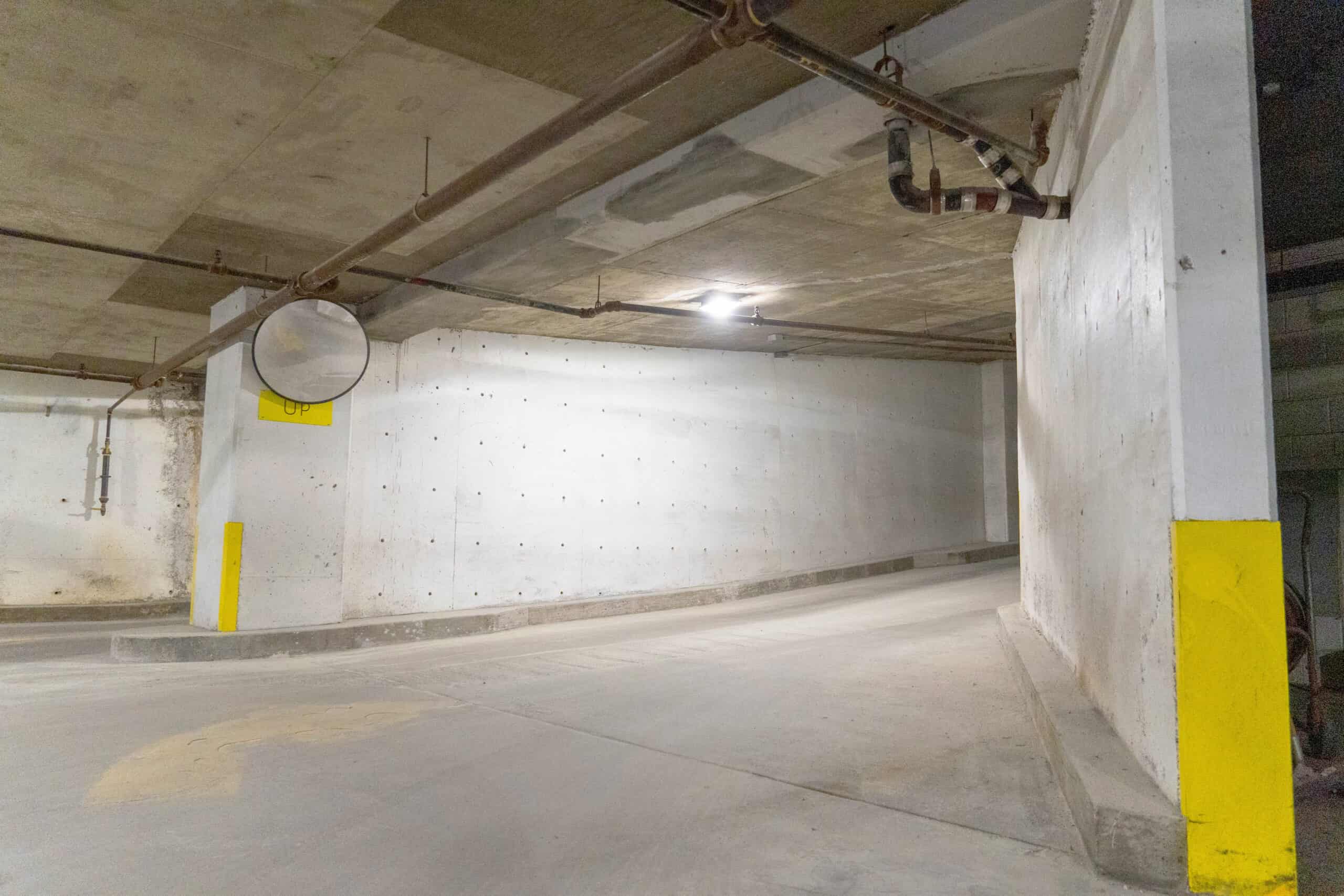 View of a two-level below-grade parking garage ramp with concrete walls and overhead piping, showing corner visibility mirror and yellow safety markings on columns.